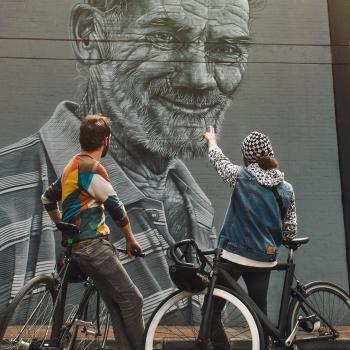 Пара на велосипедах, стоящая спиной друг к другу, смотрит на фреску колумбийского уличного искусства.