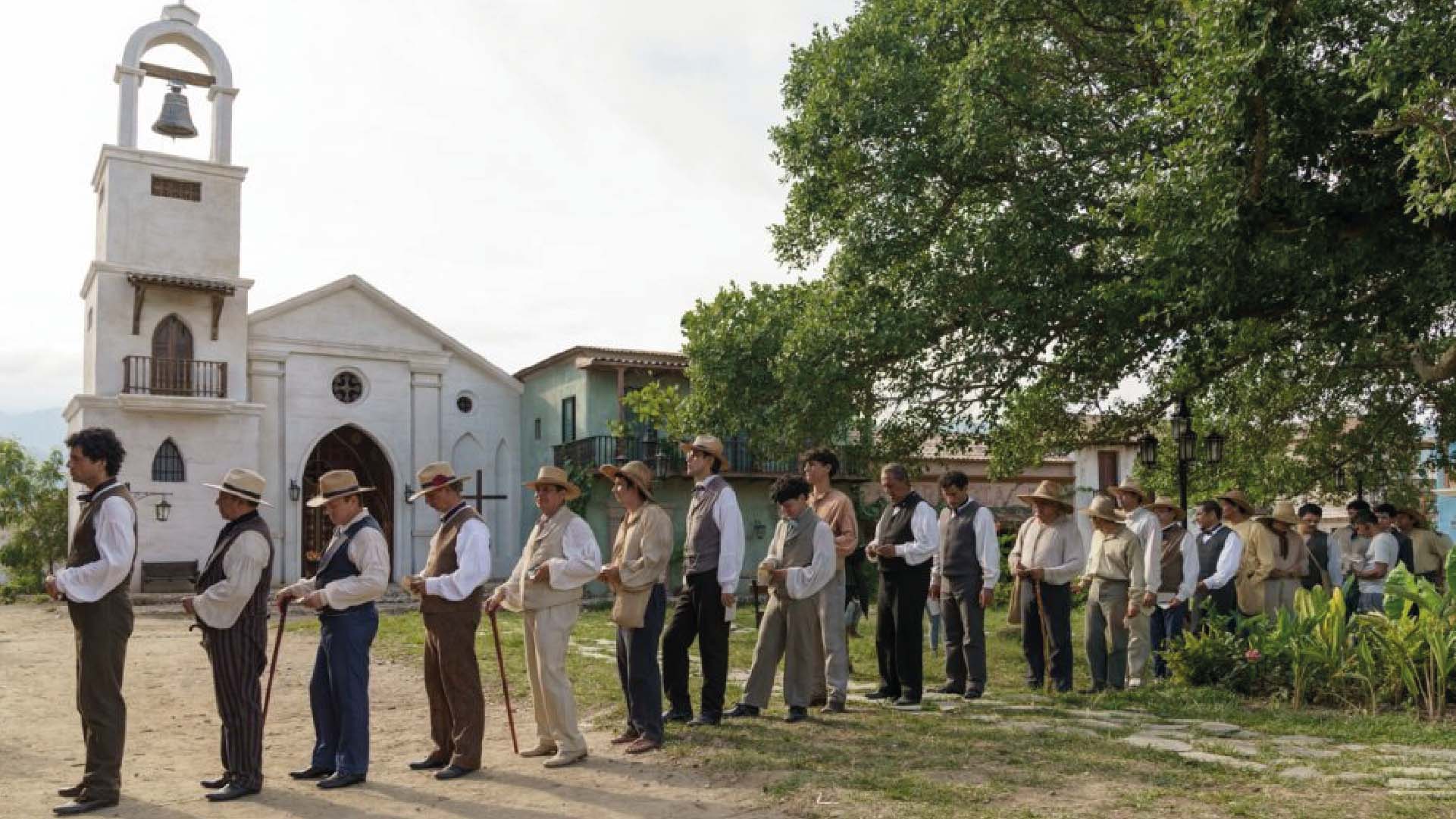 Grupo de hombres vestidos con ropa de época, usando sombreros y chalecos, hacen fila en el exterior de una iglesia blanca con campanario.