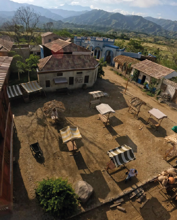 Vista aérea de un pueblo antiguo con un mercado al aire libre, donde se observan puestos de madera con techos de palma y lona rayada.