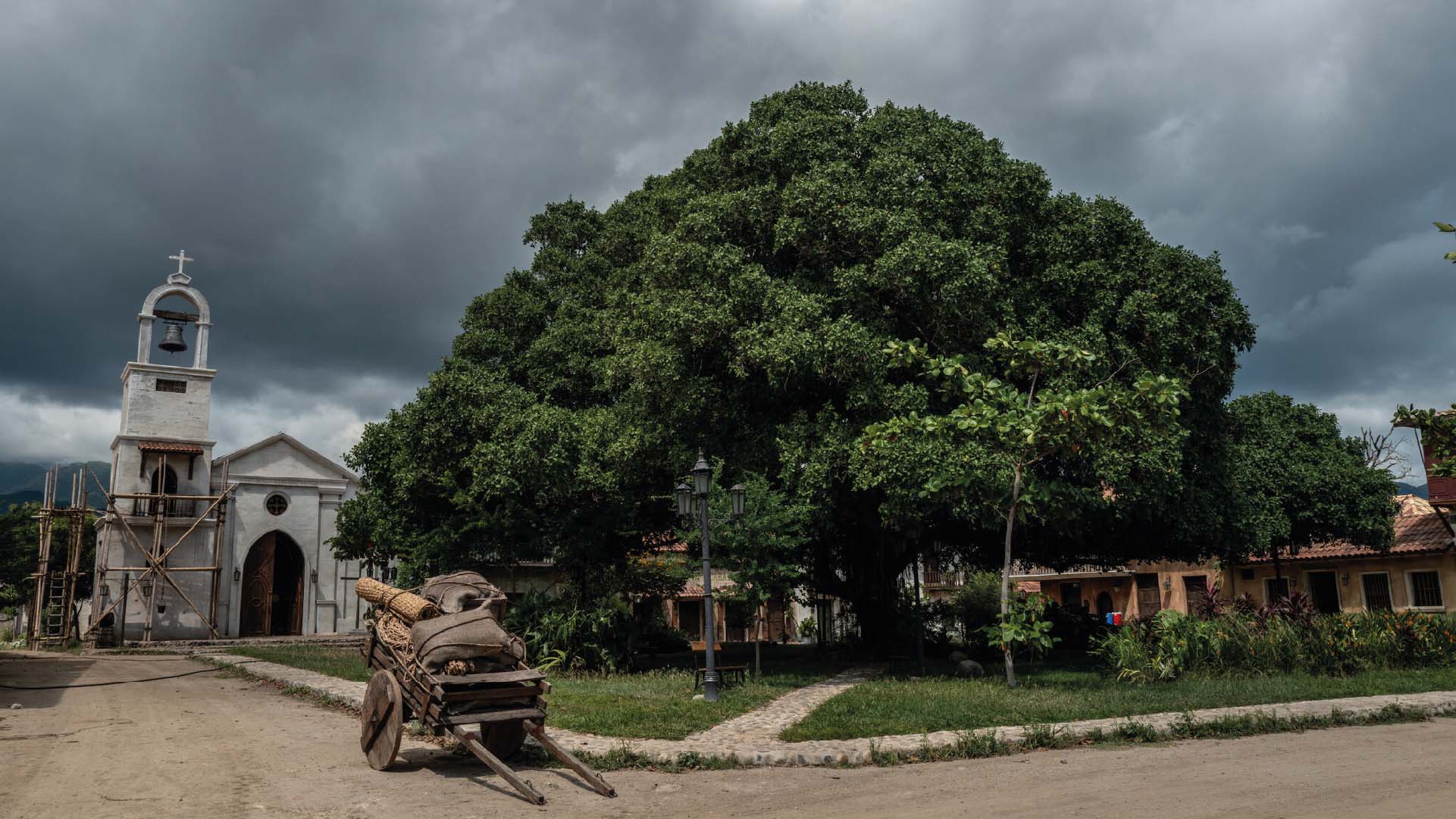 Village avec une église blanche, un grand arbre et une charrette en bois.