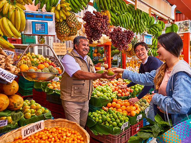 plaza de mercado llena de frutas con vendedor atendiendo a turistas extranjeros.