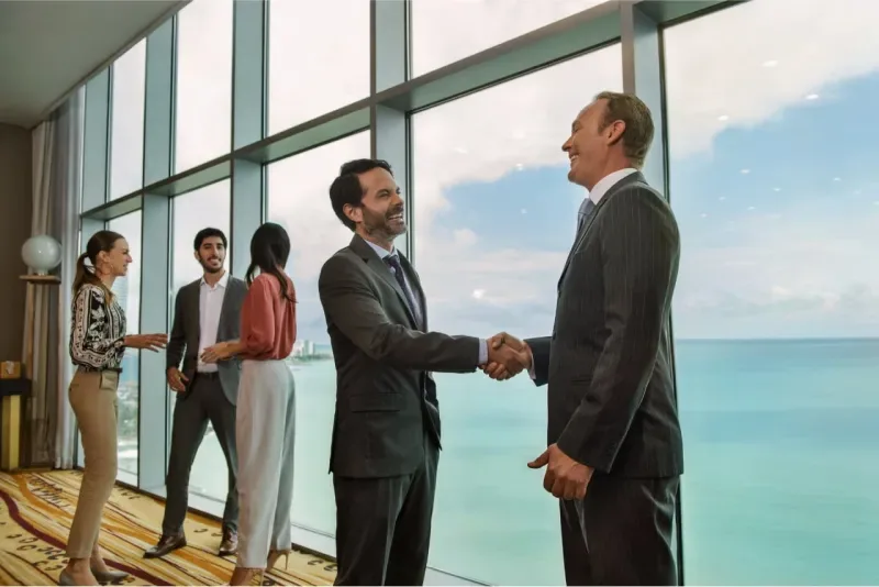 Dos hombres de negocios estrechando manos frente a una ventana panorámica con vista al mar.