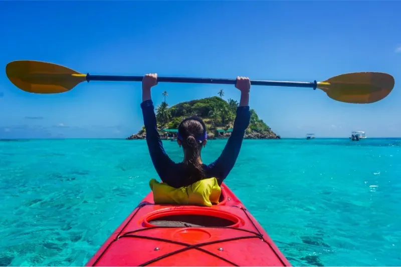 Mujer en un kayak rojo con remo levantado, en agua turquesa hacia un islote tropical.