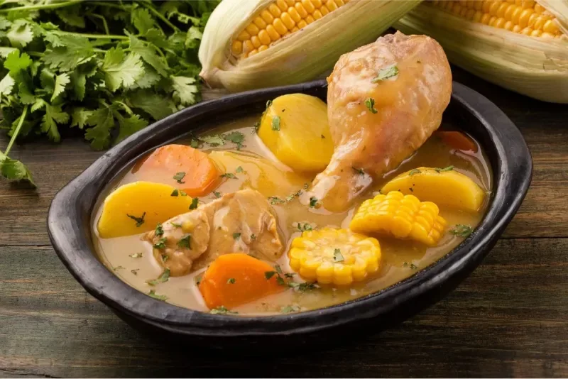 A bowl of chicken Sancocho served in a black ceramic bowl on a wooden table.