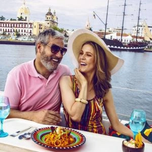 Pareja feliz comiendo en un yate, con la ciudad amurallada de Cartagena y un velero al fondo.